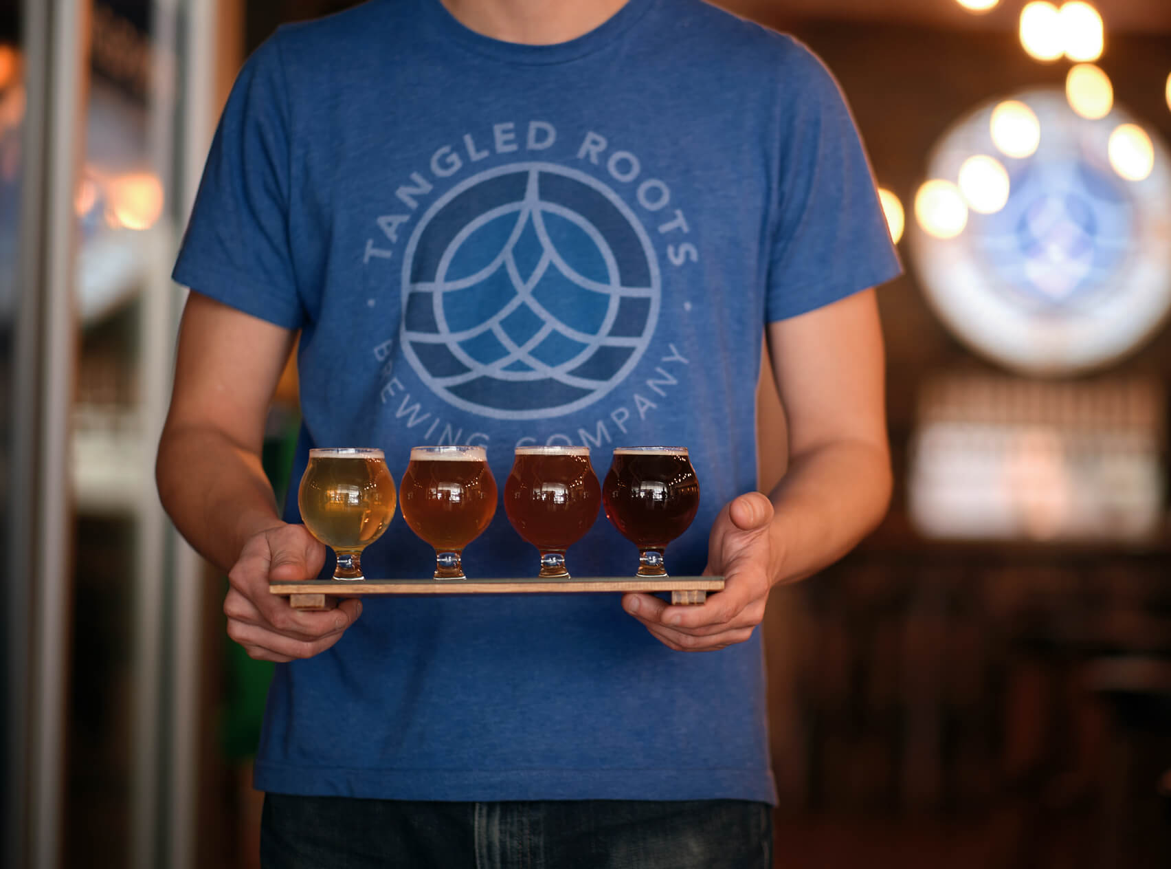 man in blue Tangled Roots Brewing Company t-shirt holding flight of four beers