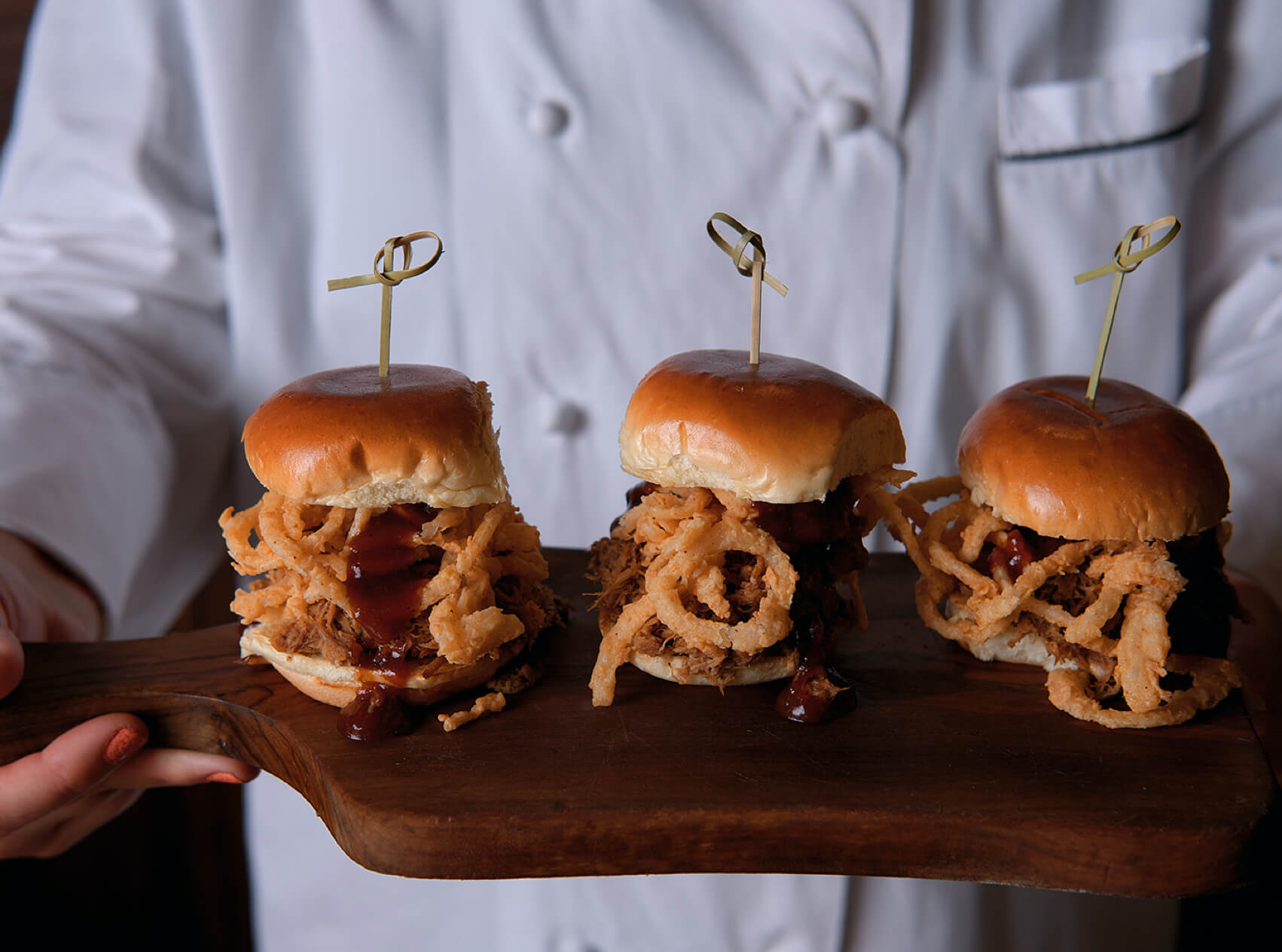 Tangled Roots chef in white chef's coat holding plate of three pork sliders with onion strings and barbeque sauce